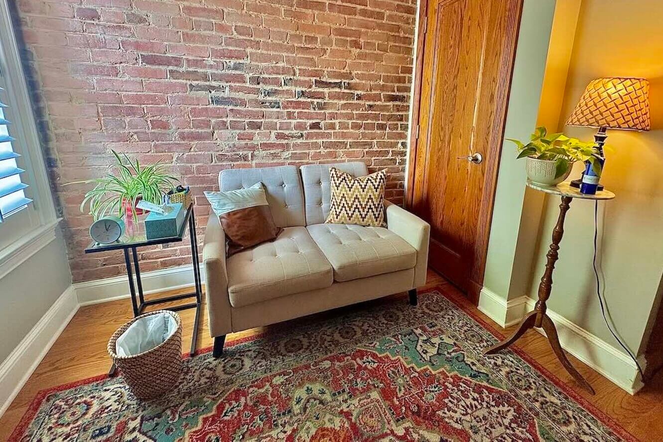 Cozy therapy office, small space therapy office with red, multicolored rug, brown couch, exposed brick and plants.